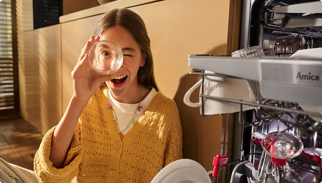 Dziewczyna przy zmywarce, wyciąga szklankę i zabawnie przykłada ją do twarzy.