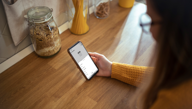 kobieta opiera się o blat w kuchni, w ręce trzyma telefon, na blacie stoi słoik z płatkami owsianymi i makaron
