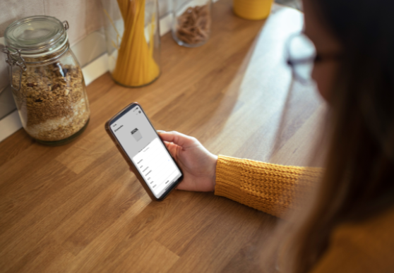 kobieta opiera się o blat w kuchni, w ręce trzyma telefon, na blacie stoi słoik z płatkami owsianymi i makaron