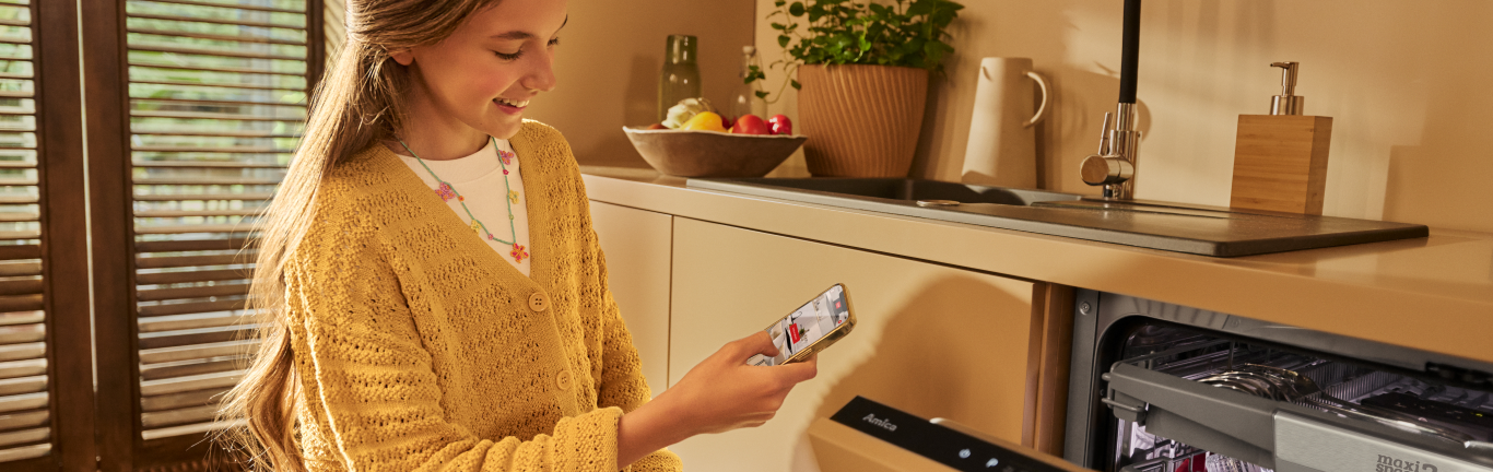 Dziewczyna przy otwartej zmywarce, z telefonem w ręce, sprawdza aplikacje